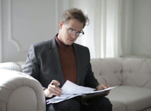 Elegant adult man in jacket and glasses looking through documents while sitting on white sofa in luxury room